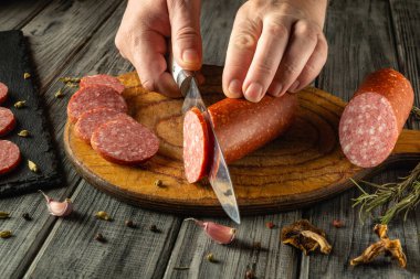 Eller ustalıkla ahşap bir tahtanın üzerinde salam dilimleri kesiyor. Sarımsak karanfilleri ve otlar, sıcak bir mutfak atmosferi oluşturur..
