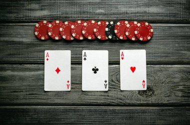 Tahta bir masada üç as kazanan bir el oluştururken, bir yığın kırmızı ve siyah poker fişi düzgünce yukarıda duruyor. Ortam rekabetçi bir eğlence akşamını çağrıştırıyor..