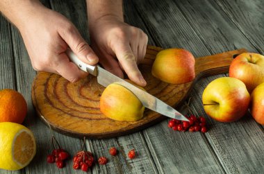 Kırsal kesim tahtasındaki sulu bir elmayı ustalıkla dilimleyen eller. Hazırlık alanı portakallar, limonlar ve kırmızı frenk üzümleri de dahil olmak üzere çeşitli taze meyvelerle çevrilidir..