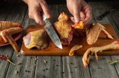 Kırsal bir mutfak ortamında, ahşap bir kesme tahtasının üzerinde çiğ tavuk parçalarını ustalıkla doğrayarak pişirmeye hazırlanıyorlar. Yakınlarda taze malzemeler var, atmosfere ekleniyor..