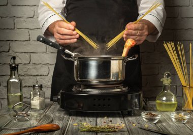 Siyah önlüklü bir şef makarna hazırlıyor, elinde kuru spagetti ve bir parça tavuğu kaynama kabının üstünde tutuyor. Taze malzemeler ve baharatlar mutfak tezgahını süslüyor..