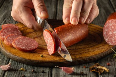 Bir insan hünerli bir şekilde sade bir tahta üzerinde sosis dilimler. Mutfakta sıcak bir atmosfer var. Taze malzemelerle gurme yemek hazırlama sanatını ön plana çıkarıyor..