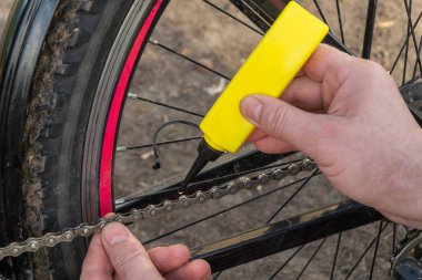 Bir kişi parkta bisikletin yanında otururken bir bisiklet zincirine kayganlaştırıcı sürüyor. Toprak kir ve güneş ışığı sıcak bir atmosfer yaratır..