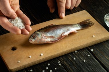 Tuzlu sazan balığı pişirmek ya da ahşap bir mutfak tahtasında Rutilus hekkelii pişirmek. Bir adamın eli çiğ balığa kaba tuz ekler..