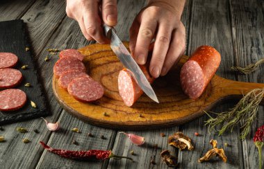 Bir insan, çeşitli baharat ve otlarla çevrili ahşap bir tahtanın üzerinde ustalıkla sosis dilimler. Sıcak mutfak atmosferi yemek pişirme deneyimini arttırır..