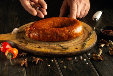 Kırsal bir mutfakta, eller ahşap bir kesim tahtasındaki büyük bir sosise baharat ekler. Hazırlığa taze otlar, sarımsak ve renkli biberler eşlik ediyor..