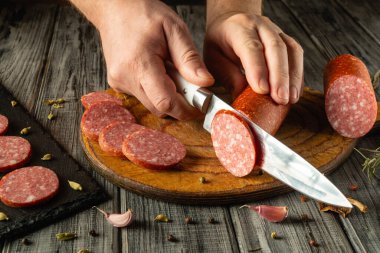 Bir çift el, tahta kesme tahtasındaki farklı salam türlerini dilimliyor. Sarımsak, karanfil ve baharatlar şarküterinin etrafını sararak, sade bir atmosfer yaratıyor..