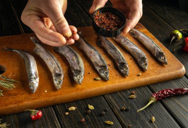 Bir insan ahşap bir tahta üzerine yerleştirilmiş taze balık sırasına nazikçe baharat serpiyor. Etrafı şifalı otlar ve çeşitli biberlerle çevrili ve sıcak bir mutfak atmosferinde..