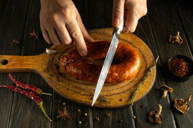 Ellerinde keskin bir bıçak var, sıcak bir mutfakta kurutulmuş baharat ve otlarla çevrili ahşap bir kesme tahtasının üzerinde dikkatlice tütsülenmiş bir sosisi kesiyor..