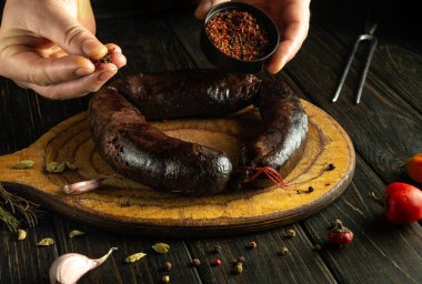 Eller, ahşap bir tahtanın üzerinde duran büyük, koyu bir sosisin üzerine ustalıkla baharat serpiyor. Hazırlık alanını çevreleyen taze malzemeler ve çatallar, sıcak bir mutfakta yemek pişirme anını ön plana çıkarıyor..