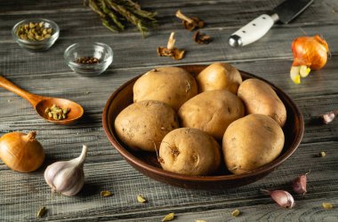 Taze patatesle dolu bir kase sarımsak, soğan ve küçük baharat kâseleriyle çevrili ahşap bir masada oturur. Malzemeler yemek hazırlığı için hazır..