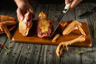 Kümes hayvanlarını ustalıkla kesen eller, ahşap bir kesim tahtasının çeşitli parçalarını sergiliyor, sıcak, kırsal bir mutfak ortamında, yanında kurutulmuş otlarla birlikte..