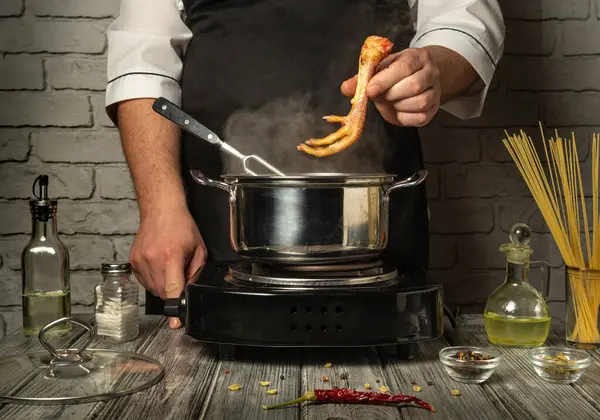 Bir şef kaynayan bir tencere çorbaya odaklanır ve dikkatlice tavuk ayağı ekler. Taze malzemeler ve baharatlar kırsal gri ahşap bir masaya serilmiştir..