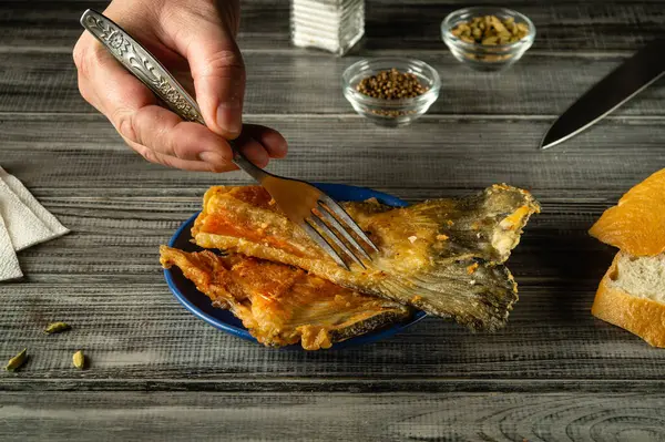 Bir el, yemek deneyimine sıcaklık katan ahşap bir masada, yanında kızarmış bir tabak altın balığın üzerinde duran bir çatalı tutar..