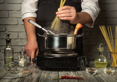 Bir şef, metal bir tencerede kaynar suya kuru spagetti ekleyerek makarna pişirmeye hazırlanır. Çevreleyen malzemeler arasında kırsal bir masada zeytinyağı, baharat ve otlar bulunur..