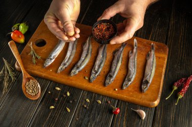 İki el ahşap kesme tahtasındaki taze temizlenmiş balığın baharatına odaklanmış durumda. Etrafa çeşitli baharatlar ve bitkiler dizilmiş, canlı bir mutfak atmosferi yaratıyorlar..