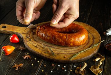 Bir insan, rengarenk baharatlar, otlar ve taze biberlerle çevrili yeni yapılmış bir sosisi ahşap bir kesme tahtasına dikkatlice bağlar ve geleneksel pişirme sürecini gözler önüne serer..