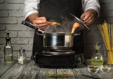 Bir şef, ocak üzerinde haşlama tenceresi hazırlar, ahşap kaşık kıpırdarken baharat ve otlar ekler. Mutfağın ahşap yüzeyli kırsal bir havası var..