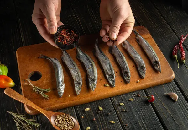 Taze balık ahşap bir kesme tahtasına yerleştirilirken, eller küçük bir kaseden baharat ekliyor. Chili biberleri, bitkiler ve domatesler hazırlık bölgesinin etrafını sararak rahat bir atmosfer yaratıyor..