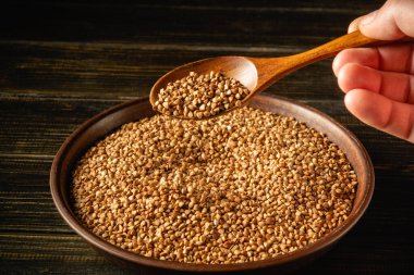 Karabuğday taneleriyle dolu bir kasenin üzerinde tahta bir kaşık tutan bir el. Sahne, tanelerin dokusunu ve rengini aktararak doğal görünümlerini vurgular..