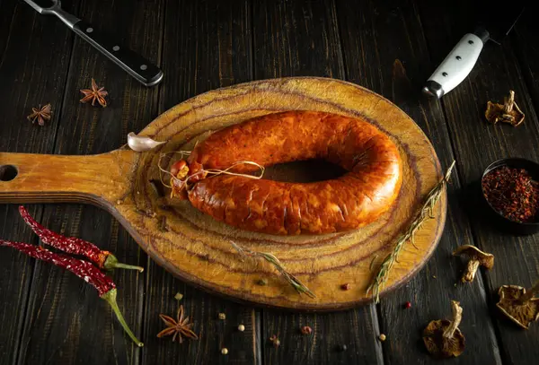 Büyük, el yapımı bir sosis ahşap bir kesme tahtasının üzerinde duruyor. Etrafını çeşitli kurutulmuş baharatlar ve otlar sararak kırsal mutfak atmosferini geliştirir. Doğal ışık sahneyi aydınlatır..