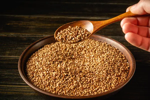 Karabuğday taneleriyle dolu bir kasenin üzerinde tahta bir kaşık tutan bir el. Sahne, tanelerin dokusunu ve rengini aktararak doğal görünümlerini vurgular..