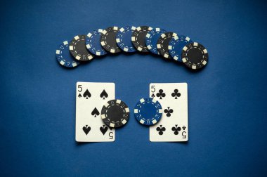 Bir kart oyununda birkaç poker fişi tarafından çevrelenmiş iki beş kart sergileniyor. Mavi arka plan oyun unsurlarına zıtlık katarak onları akşam atmosferinde öne çıkarıyor..