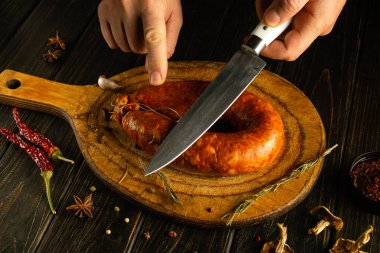 Bir insan ustalıkla bir parça zanaatkar sosisi tahta bir kesme tahtasıyla keser. Çevrelerinde acı biber ve şifalı bitkiler de dahil olmak üzere çeşitli baharatlar var, kırsal bir mutfak ortamı yaratıyorlar..