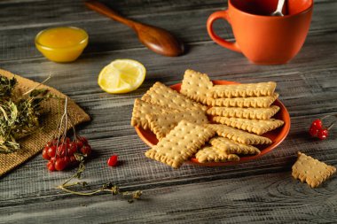 Bir tabak gevrek bisküvi bir fincan çay, limon dilimleri ve küçük bir kase bal eşliğinde ahşap bir masada oturur. Taze böğürtlen ve şifalı bitkiler bu harika düzeni tamamlar..