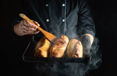 Bir fırıncı bir tepsi taze pişmiş ekmek sunar, ahşap bir spatula tutarken buhar salıyor, karanlık bir mutfak ortamında aşçılık becerilerini sergiliyor..