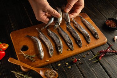 Elleri ahşap bir kesme tahtasına yerleştirirken birkaç taze balık tutuyor. Çeşitli otlar ve baharatlar sebzelerle birlikte balıklara eşlik ederek canlı bir mutfak görüntüsü yaratıyor..