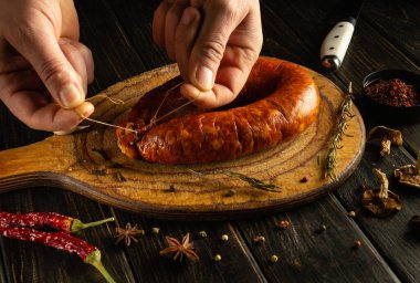 Eller ahşap bir tahtanın üzerinde, baharatlar ve kurutulmuş otlarla çevrili ev yapımı sosisi ustalıkla hazırlar, sıcak bir mutfak atmosferinde geleneksel mutfak sanatının özünü yakalar..
