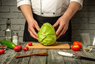 Bir şefin elleri büyük bir lahana kafasını ahşap bir kesme tahtasının üzerinde tutar. Domatesler, salatalıklar ve turplar çevreyi sararak canlı bir atmosfer yaratırlar..