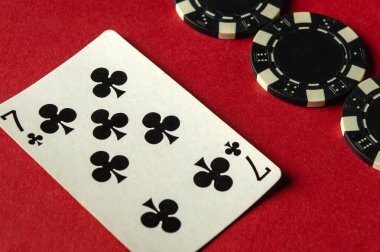 Sinek yedili bir kart kırmızı bir oyun masasının üzerinde duruyor. Birkaç yuvarlak siyah-beyaz poker fişinin yanında. Oyuncular canlı bir akşam poker oyununa girişiyor..