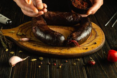 Eller, sarımsak, otlar ve acı biberle bezenmiş ahşap servis tahtasının üzerinde duran koyu renkli sosislerin üzerine baharat serpiyor. Hava sıcak ve davetkar..