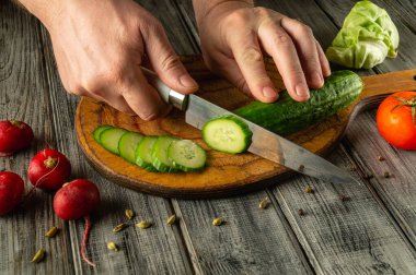 Bir insan ustalıkla taze bir salatalığı ahşap bir kesme tahtasıyla ince dilimlere ayırır. Yakınlarda, parlak kırmızı turplar ve bir domates yatar, kırsal mutfak düzenine renk katar..