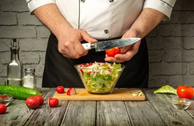 Bir şef domatesleri marul, salatalık ve turpla dolu canlı bir salatayı rahat bir mutfakta ahşap bir tezgahta ustalıkla doğruyor..