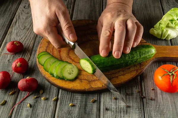 Elleri ustalıkla yeni biçilmiş bir salatalığı ince dilimlere ayırıyor. Ahşap kesme tahtasının etrafında canlı domatesler, turplar ve marul var. Bu da yemek pişirme atmosferi yaratıyor..