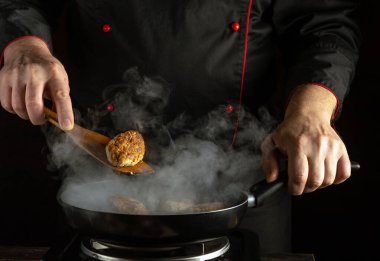 Becerikli bir şef, buhar ve dumanla çevrili bir tavada köfte hazırlar. Aşçı yemek pişirme sürecine odaklanırken, sıcak aydınlatma atmosferi arttırır..