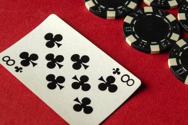 Yakın çekim görüntüsünde, oyun atmosferini yansıtan kırmızı bir masada, birkaç siyah kumarhane fişi yanına yerleştirilmiş bir kulüp kartı yer alıyor..