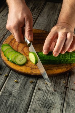 Eller ustalıkla taze bir salatalığı ahşap bir kesme tahtasındaki dilimlere ayırdı. Güneş ışığı, mutfakta sıcak ve davetkar bir atmosfer yaratıyor..