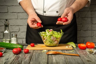 Siyah önlüklü bir şef, iyi aydınlatılmış bir mutfaktaki ahşap bir masadaki karışık yeşilliklere turp ekleyerek taze bir salata hazırlıyor..