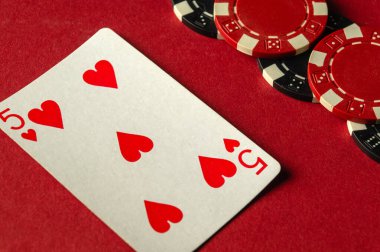 Kupa beşlisi, çeşitli poker fişlerin yanında kırmızı bir masaya uzanır. Arkadaşları arasında strateji ve rekabetle dolu bir oyun gecesinin heyecanını yakalar..