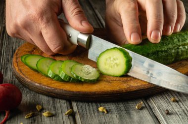 Elleri ustalıkla taze bir salatalığı tahta bir kesme tahtasıyla doğruyor. Parlak yeşil dilimler sağlıklı yemek pişirmeyi vurgulayan salatalık ve diğer taze sebzelerin yanına güzelce yerleştirilir..