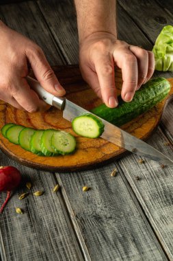 Elleri ustalıkla bir ahşap kesim tahtasında salatalık dilimler, turp ve marul ile çevrili, kırsal bir mutfak ortamında yemek hazırlama sanatını sergiler..