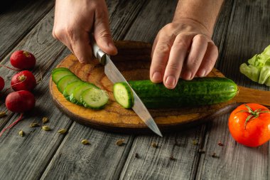 Elleri ustalıkla bir salatalığı, turp, marul ve domates gibi taze sebzelerle çevrili ahşap bir tahtanın üzerinde doğruyor..