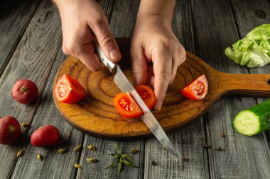 Elleri ustalıkla olgunlaşmış domatesleri otlar ve diğer sebzelerle çevrili ahşap bir kesme tahtasının üzerinde dilimliyor. Sıcak atmosfer sıcak bir mutfak titreşimi sergiliyor..