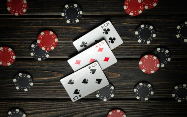 Üç beşli bir iskambil kağıdı seti, çeşitli poker fişleri ile çevrili ahşap bir masanın üzerinde duruyor. Ilık ışıklandırma, arkadaşların oyun oynamaları için rahat bir ortam oluşturur..