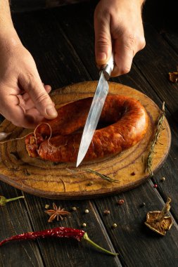 Eller ustalıkla büyük, sulu bir sosisi kesme tahtasının üzerinde dilimler. Çevreleyen malzemeler arasında bitkiler, baharatlar ve biberler var. Mutfak atmosferine kırsal bir dokunuş ekleniyor..