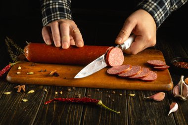 Eller, taze otlar, baharatlar ve sarımsak ile çevrili ahşap kesme tahtasındaki ev yapımı sosis kütüğünü ustalıkla dilimliyor..
